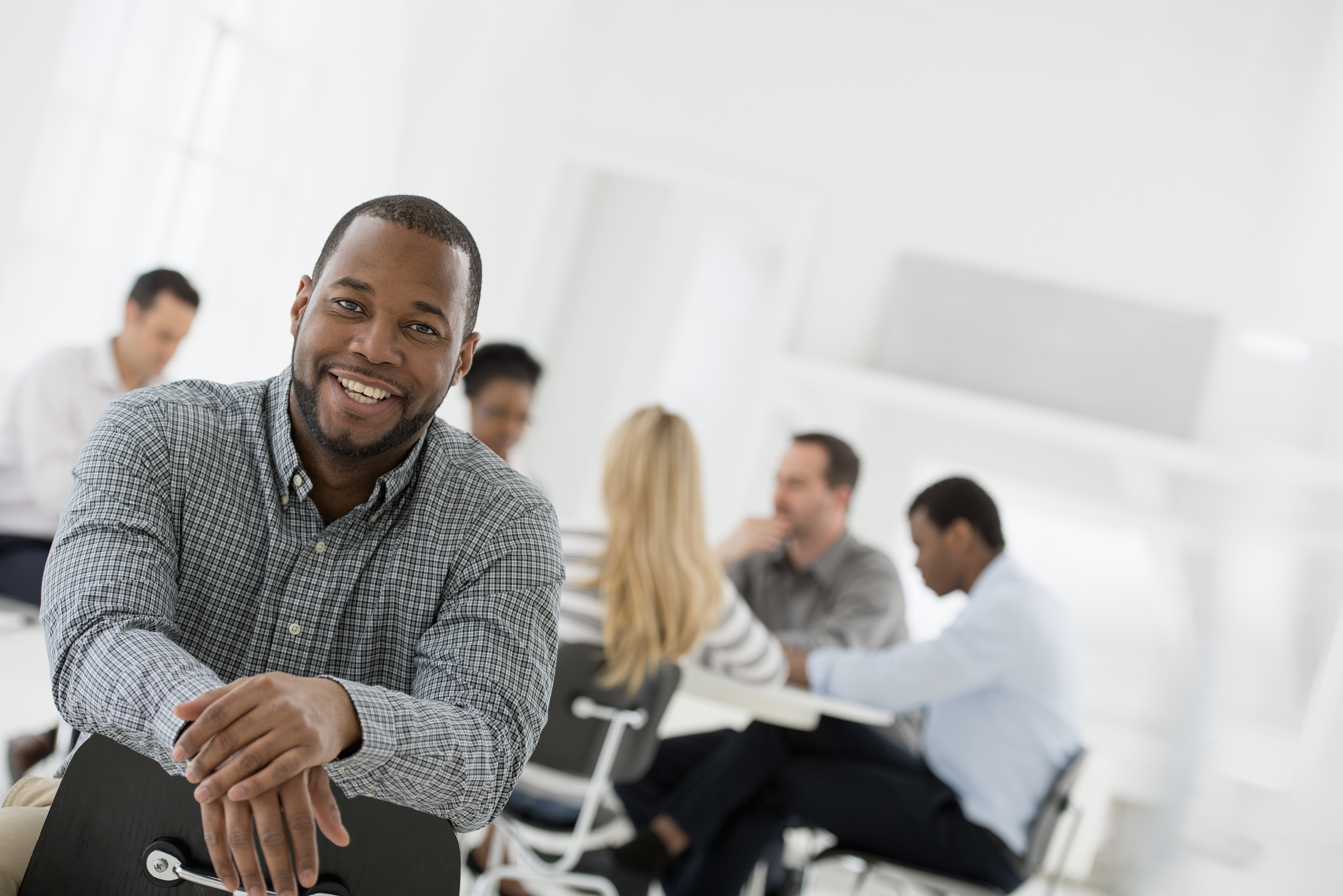 Featured image: Smiling man leaning on the back of a chair in the foreground, looking at the camera, with coworkers in the background seated around a table in a bright office meeting room. - Read full post: What to Do When Your Career Feels Stuck