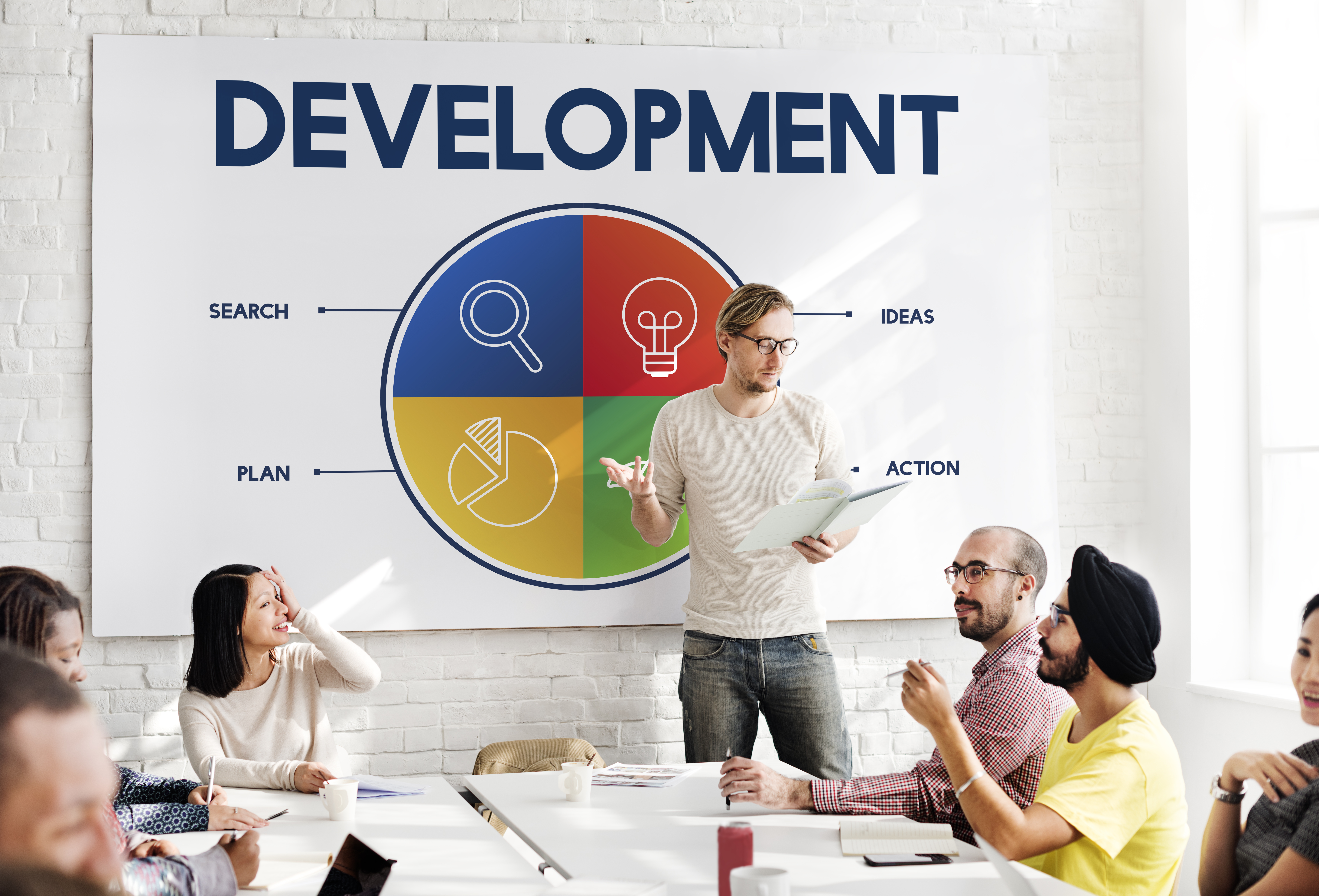 Featured image: A group of colleagues in a meeting room discussing ideas while a presenter stands in front of a wall graphic titled “Development,” showing a circular diagram with sections for search, ideas, plan, and action. - Read full post: Career Development: How to Recognise Growth at Work