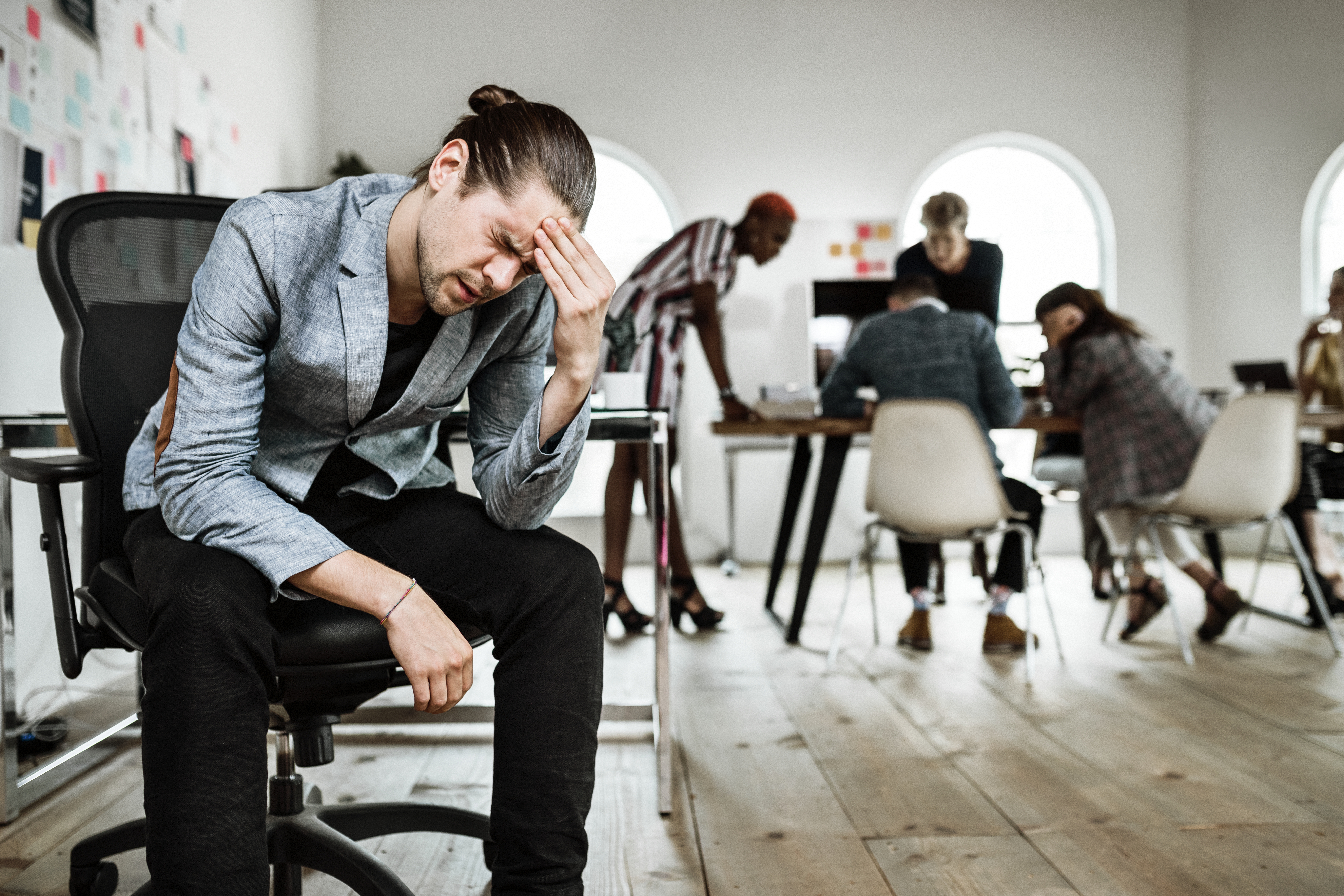 Featured image: A stressed man sits on an office chair holding his head, while colleagues work and talk in the background of a busy open-plan office. - Read full post: Mental Health at Work: What It Really Feels Like Day to Day