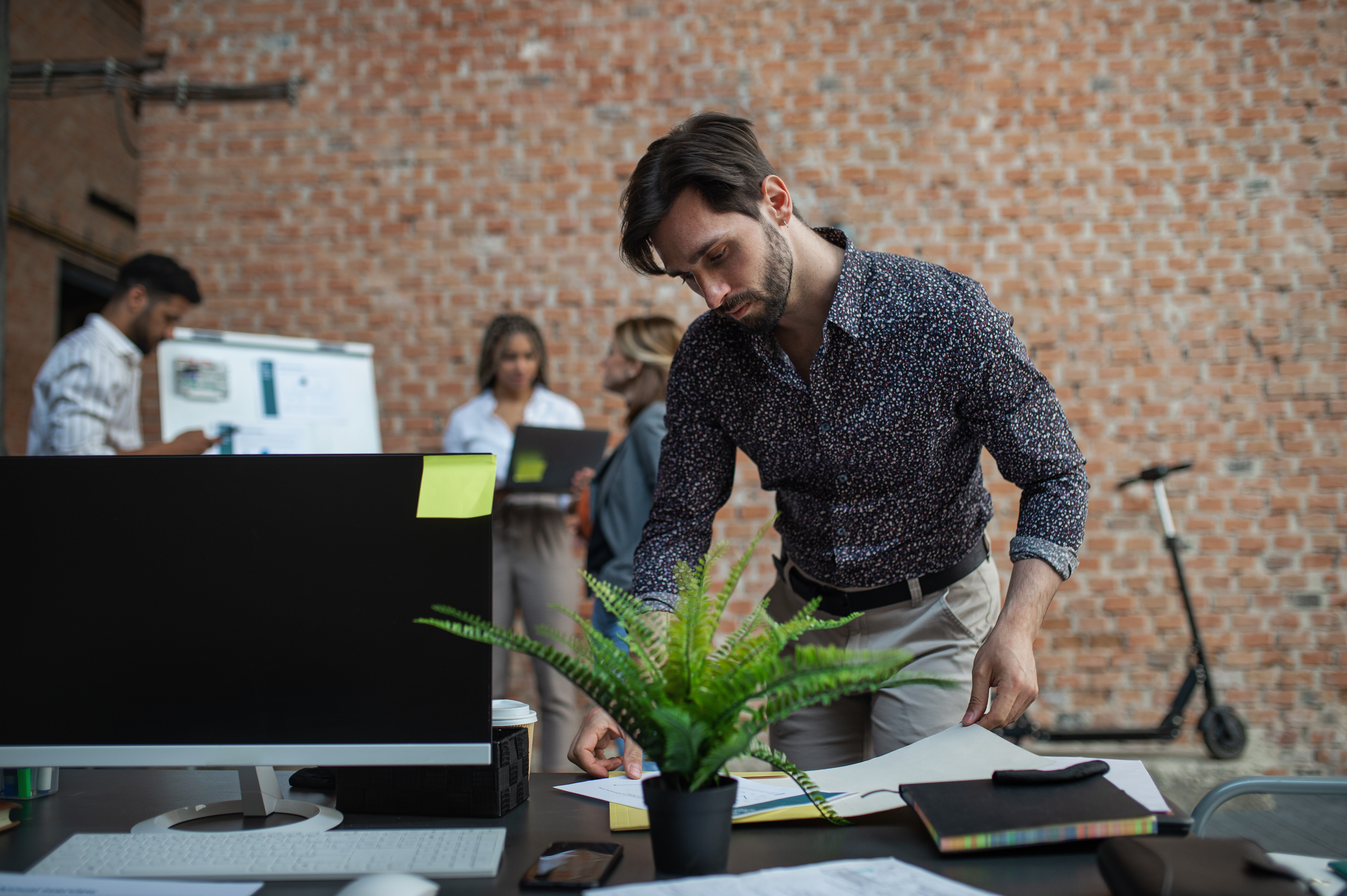Featured image: A man stands at a desk arranging documents beside a computer and potted plant in a modern office, with colleagues working in the background. - Read full post: Growing at Work Without Burning Out