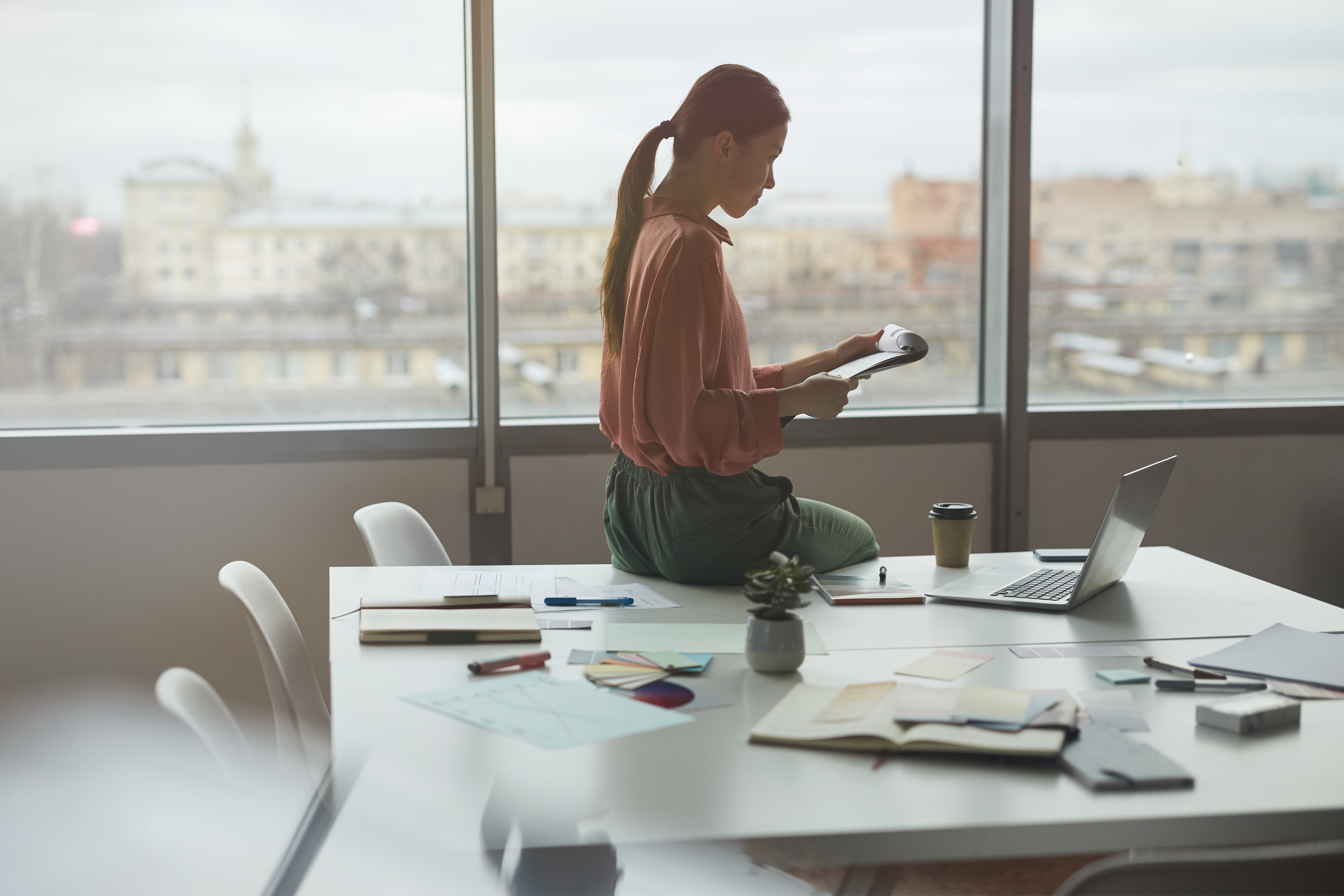Featured image: A woman sits on a desk in a quiet office, reviewing papers beside a laptop, notebooks, and coffee, with large windows overlooking a city. - Read full post: Resilience at Work: Why Slowing Down Can Help You Move Forward