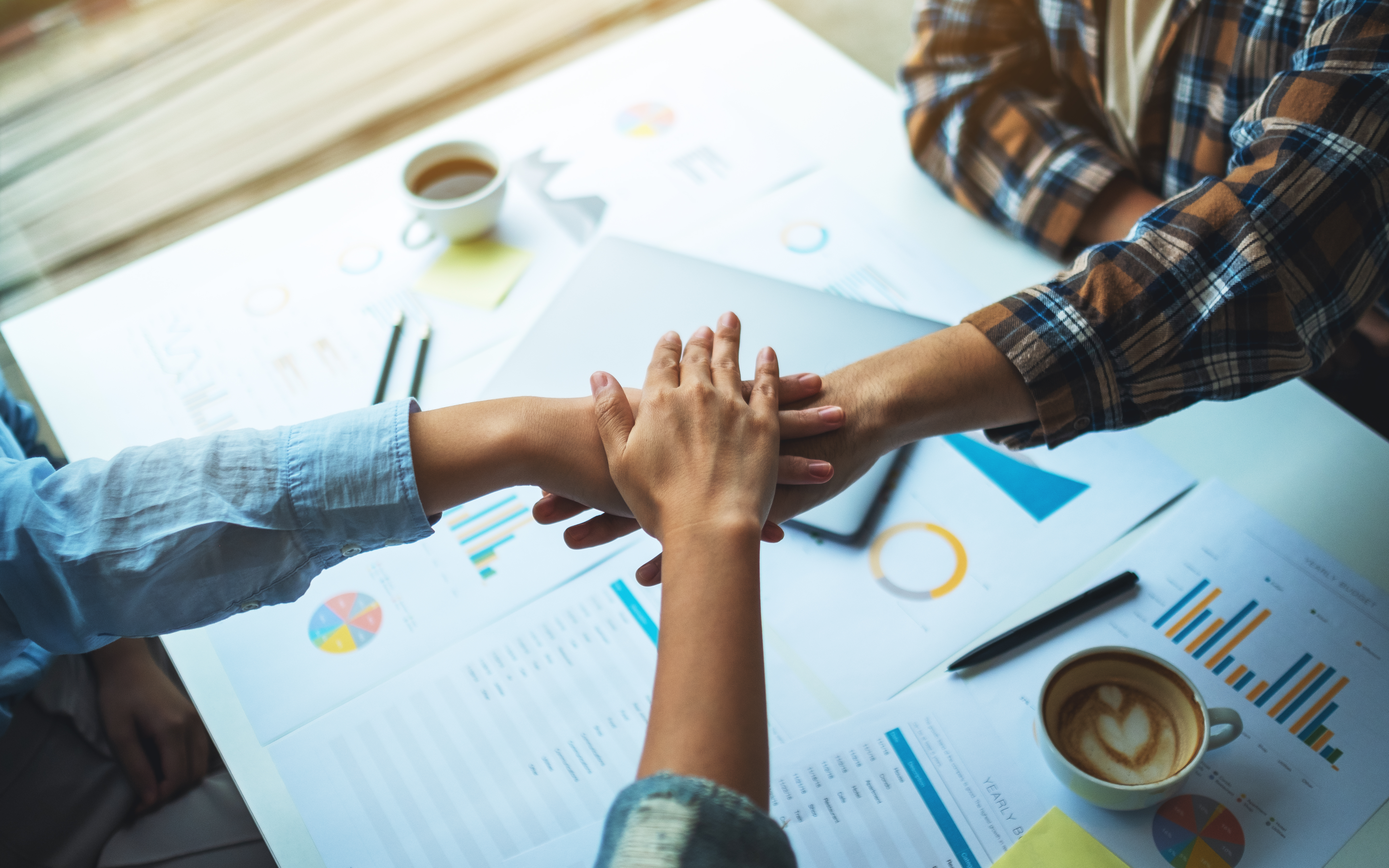 Featured image: Three people stacking their hands in a gesture of unity and teamwork over a white desk cluttered with colorful business charts, pie graphs, bar graphs, a silver laptop, a white coffee mug, yellow sticky note, black pens, and scattered papers in a well-lit office setting. - Read full post: Workplace Communication: Why Clear Conversations Matter During Late Summer