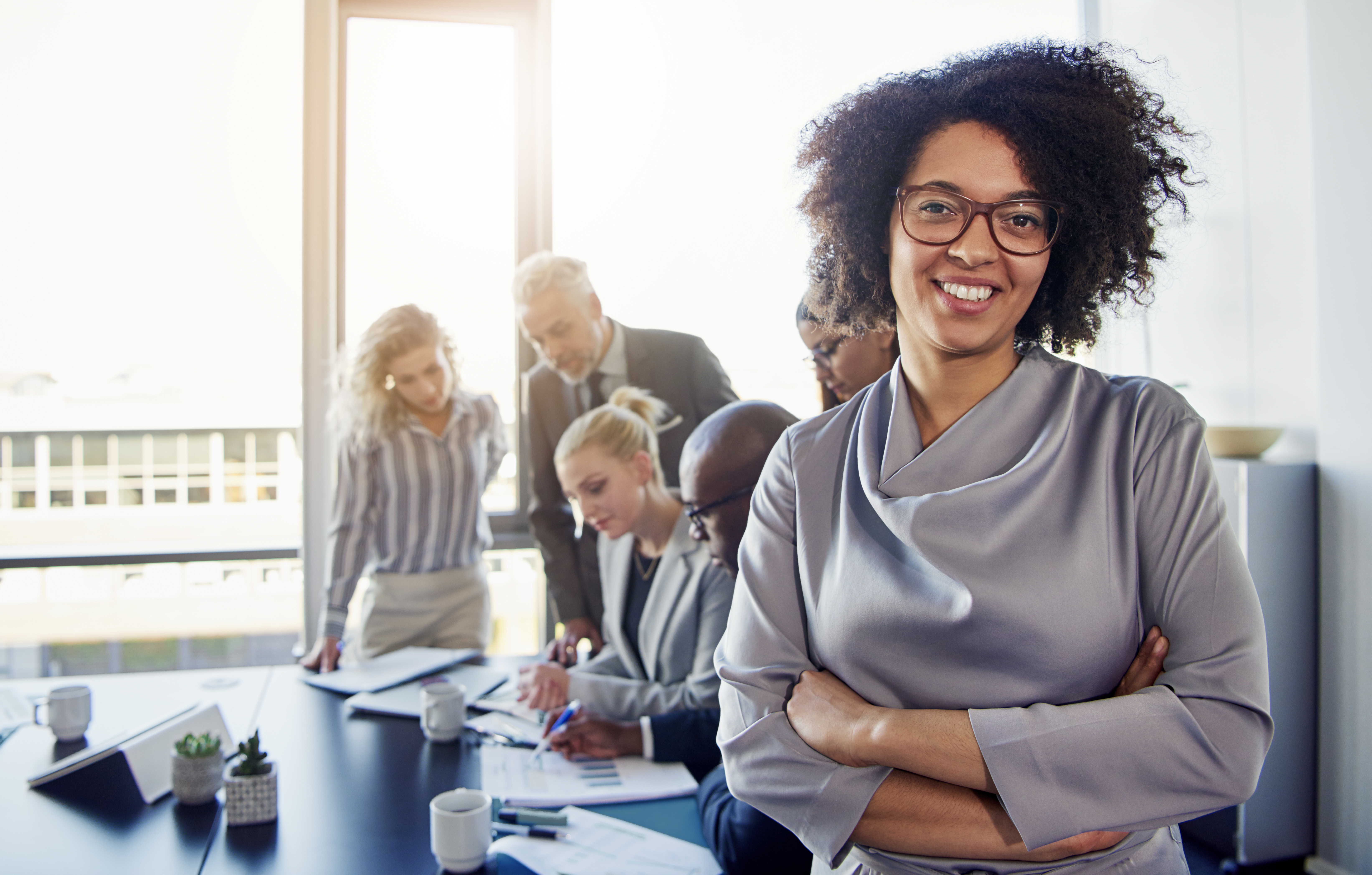 Featured image: A confident woman standing with arms crossed in a bright office, smiling at the camera while colleagues collaborate at a meeting table in the background. - Read full post: Authenticity at Work: Setting a Sustainable Tone for the New Year