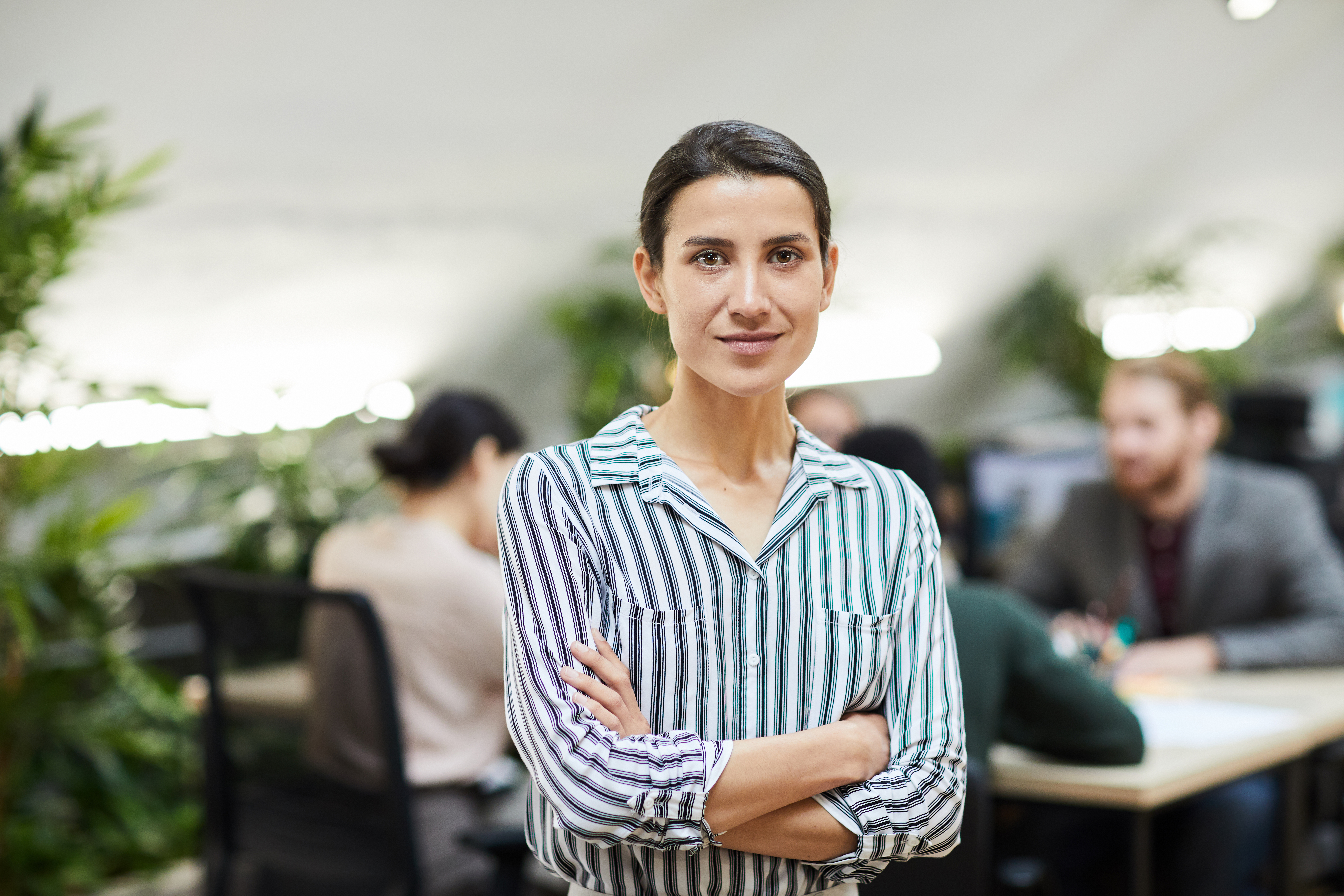 Featured image: Woman standing with arms crossed in a modern open-plan office, looking at the camera, with coworkers working at desks in the background and plants around the workspace. - Read full post: Resilience at Work: What Sustained Pressure Reveals About Your Organisation