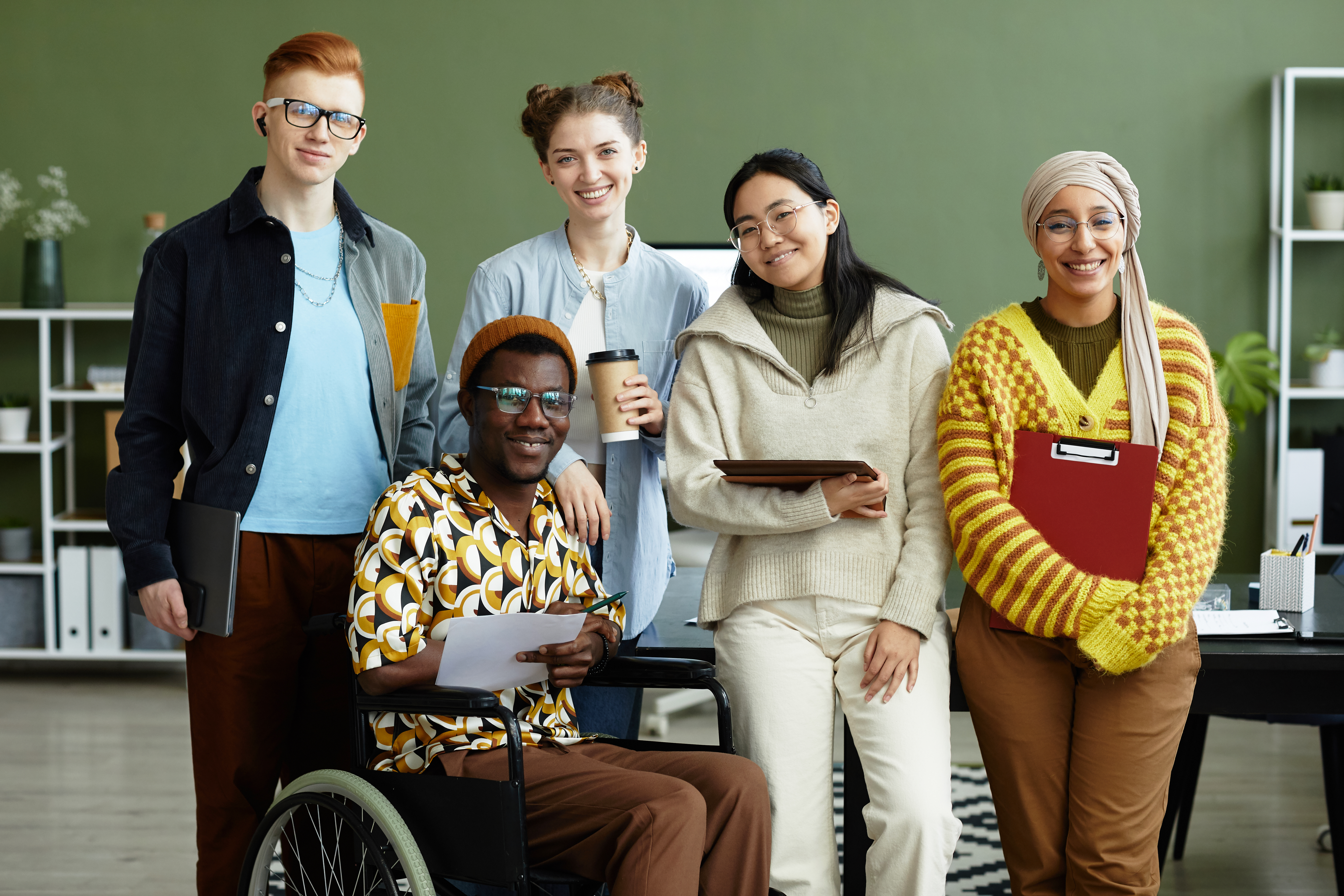 Featured image: Five coworkers of diverse backgrounds pose together in an office, smiling at the camera, with one person seated in a wheelchair holding papers while others hold a tablet, notebook, clipboard, and coffee cup. - Read full post: Belonging at Work: Why It Matters and How HR Can Strengthen It
