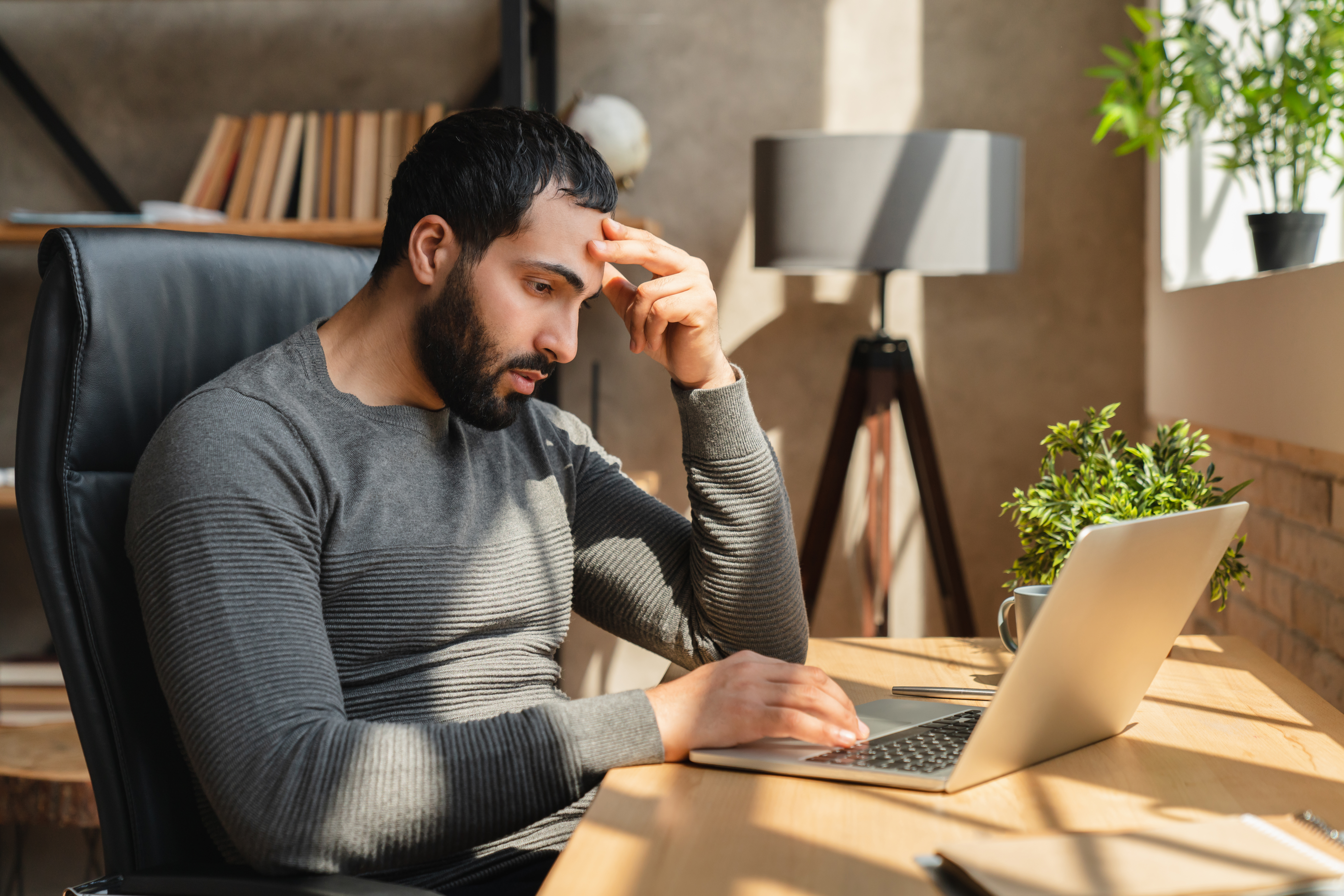 Featured image: A man sits at a desk working on a laptop, resting his hand on his forehead and looking tired or stressed in a home office with plants and natural light. - Read full post: Men’s Mental Health at Work