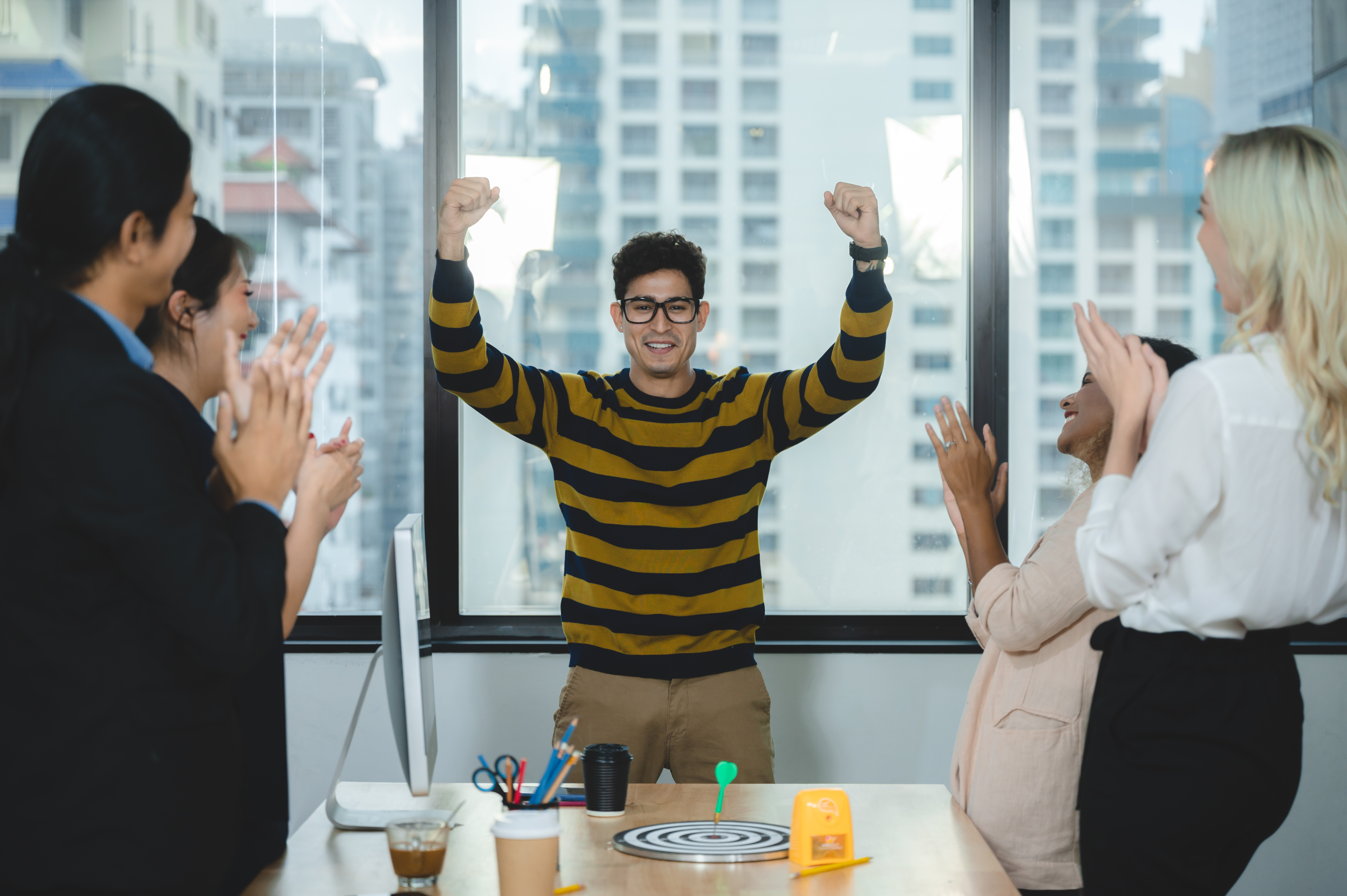 Featured image: Man standing at a desk with both arms raised in celebration while coworkers around him clap and smile, inside a bright office with large windows and city buildings visible outside. - Read full post: How High Performance Actually Holds Up